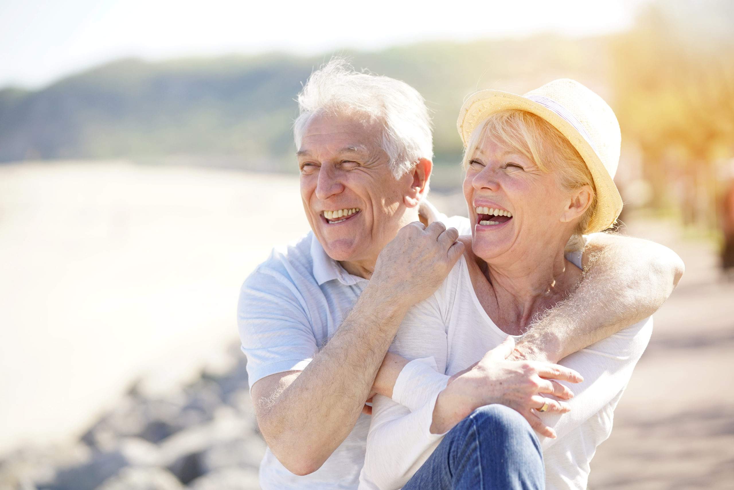 An older couple laughing together outdoors by a waterside on a sunny day.