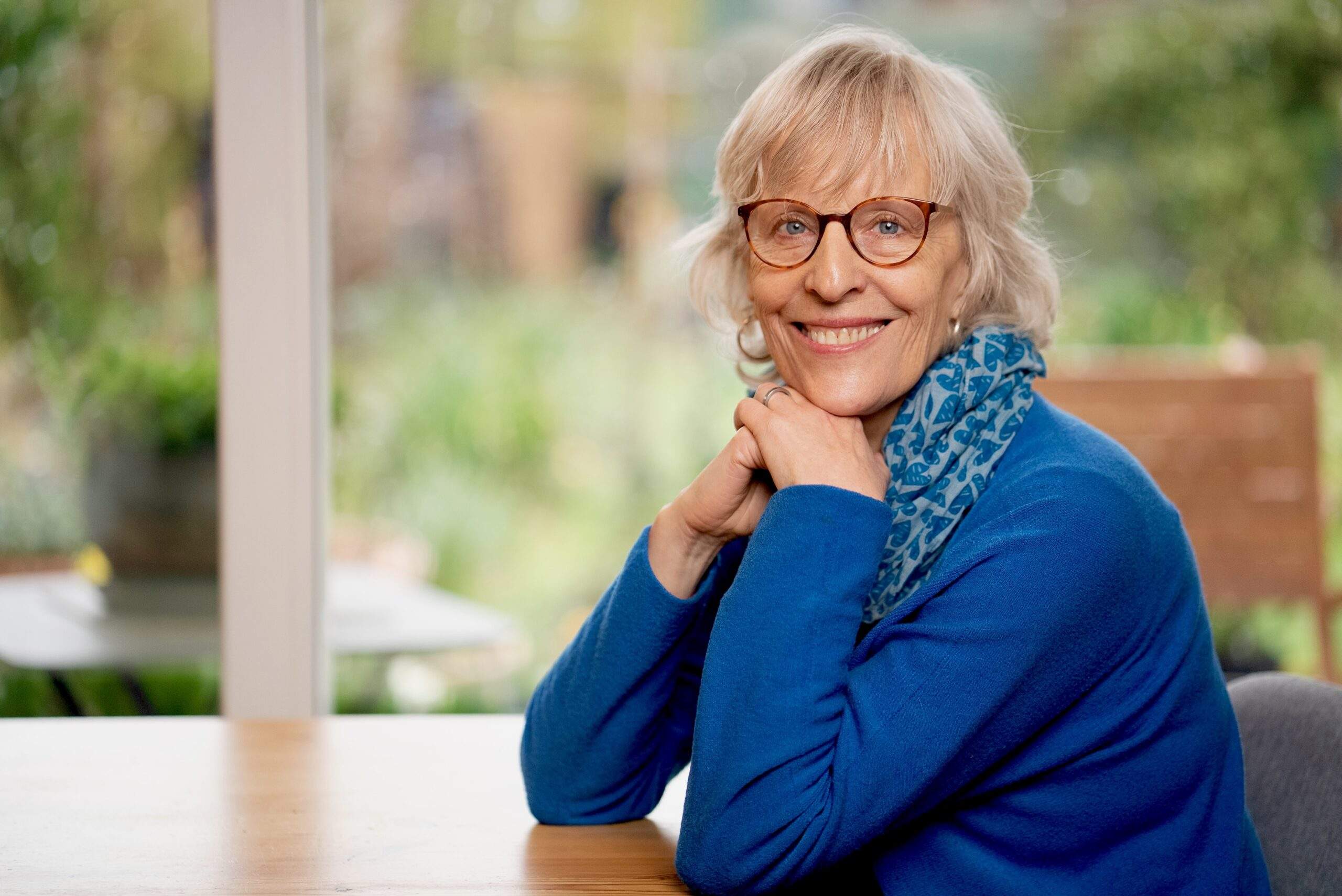 Vidyamala Burch, an older woman wearing a blue shirt and scarf, with shoulder-length hair and a vibrant smile, sits with her elbows on a table and her chin resting on her hands.