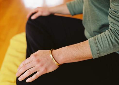A person with a golden bracelet on their wrist sits cross-legged on a yellow cushion. The individual is wearing a long-sleeved green top and black pants. Their hands rest on their knees, and the wooden floor is partially visible in the background.