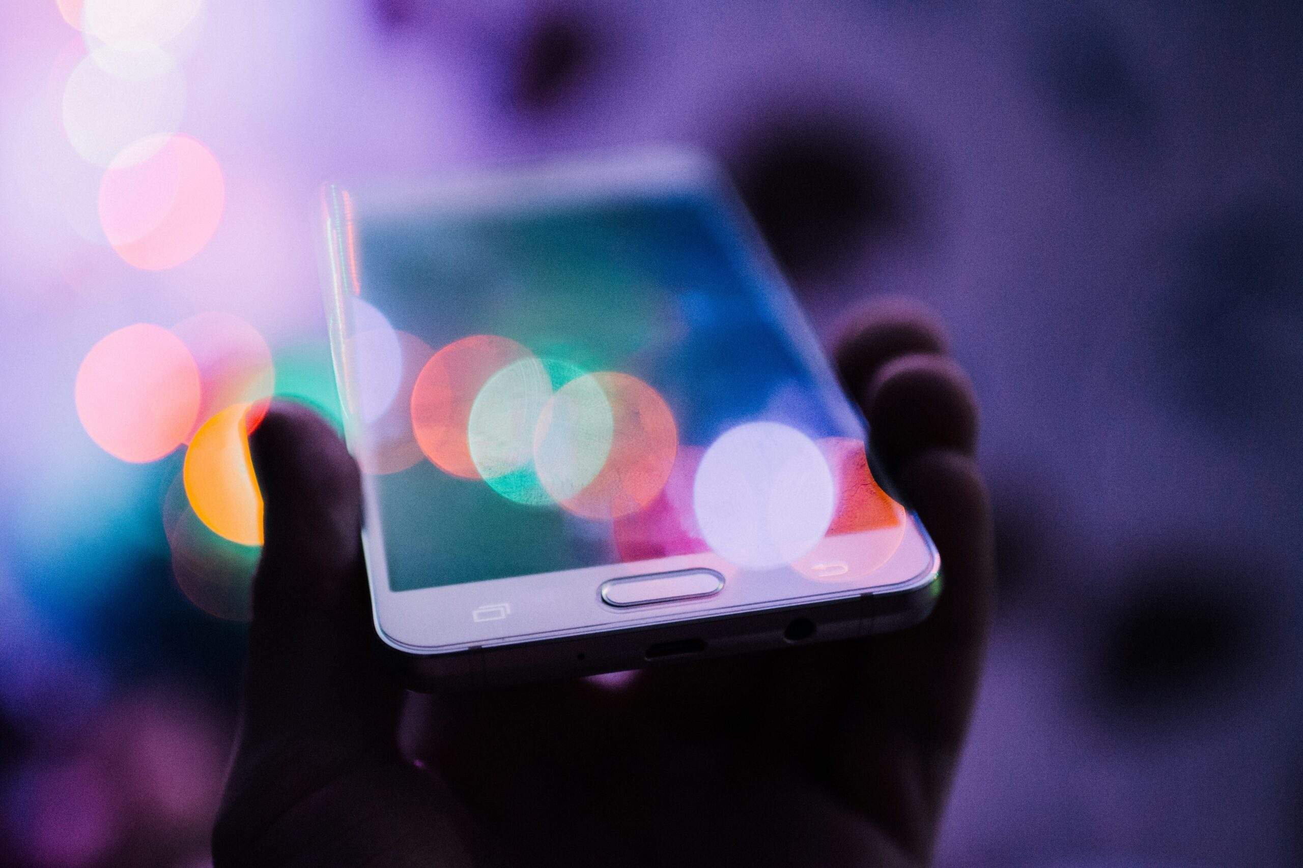 A hand holding a smartphone with colorful, blurred lights in the background, reminding you to pause before you post on social media.