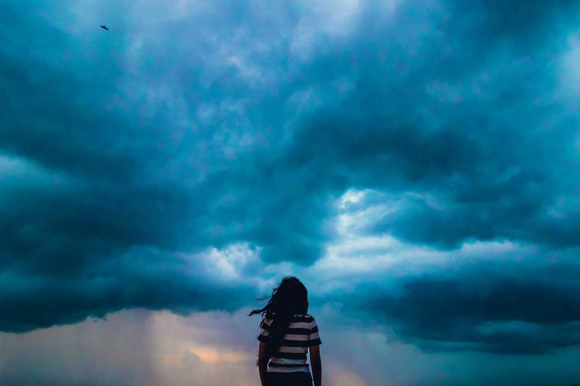 Person in striped shirt stands under dramatic, cloudy sky, sunlight breaking through—a visual echo of emerging from vicarious trauma.