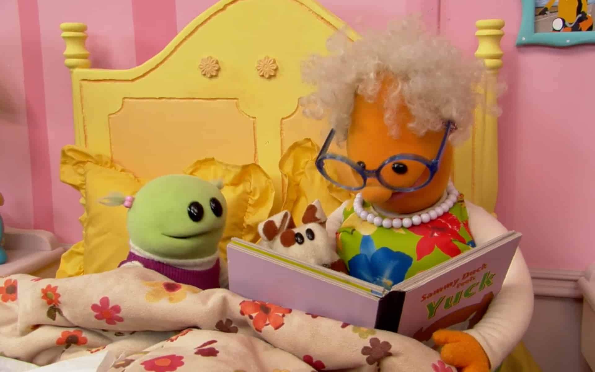 Two stuffed animals sit on a bed reading a picture book together in a child's bedroom.