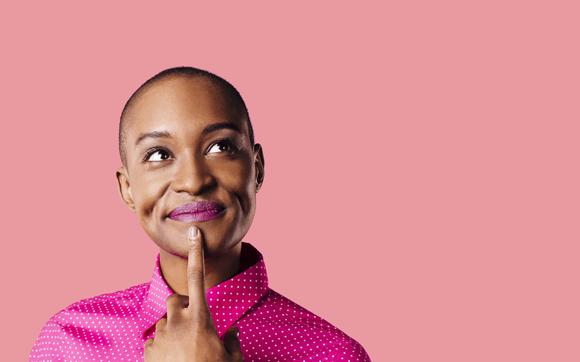 A 12-Minute Loving-Kindness Practice to Cultivate Curiosity—A Black woman wearing a pink shirt has a thoughtful expression on her face with one finger resting on her chin in front of a pink studio background.