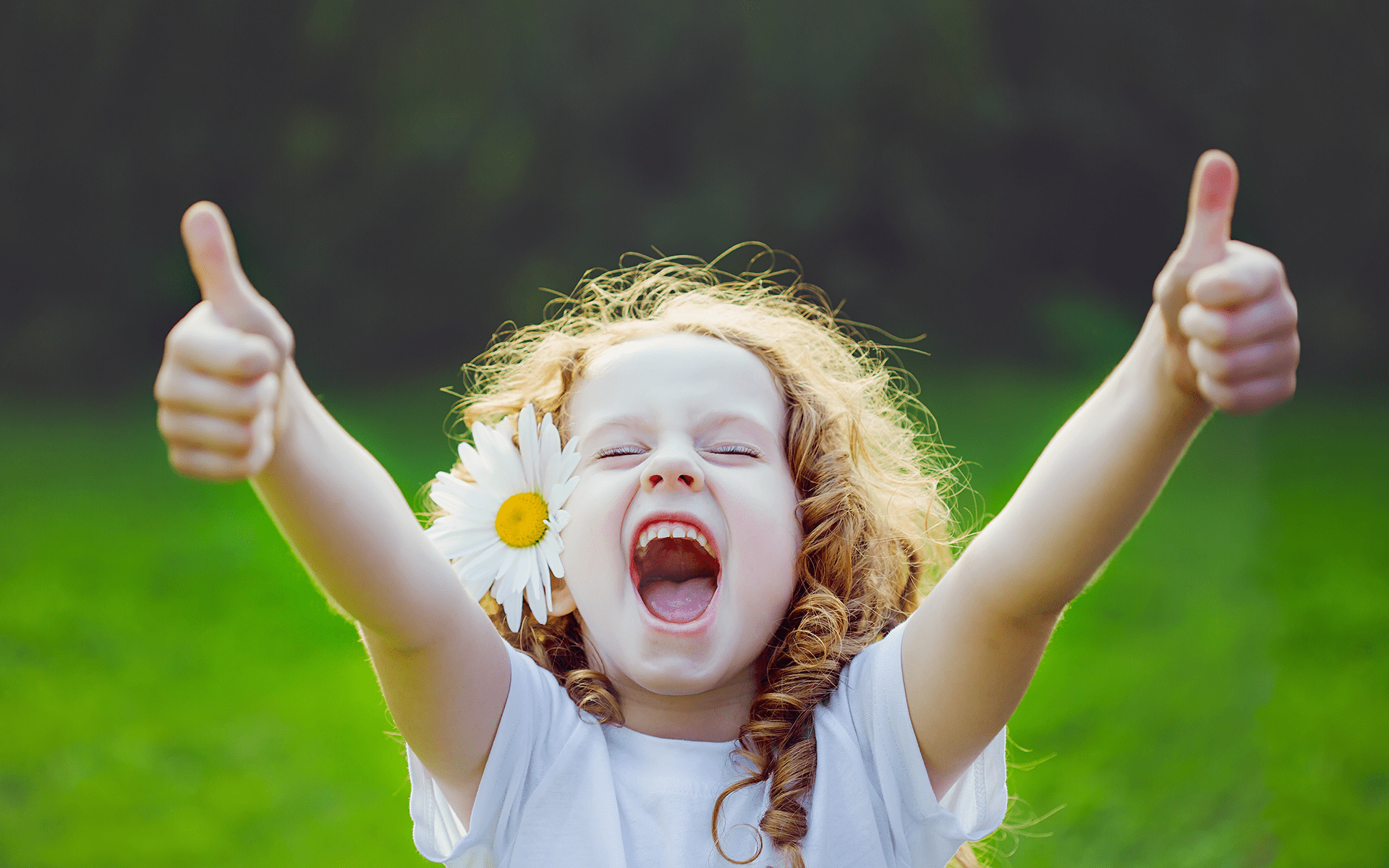 Photo of a little girl with light skin and red curly hair standing outside with a daisy tucked behind her ear with a big smile and giving two thumbs up to the camera. joy
