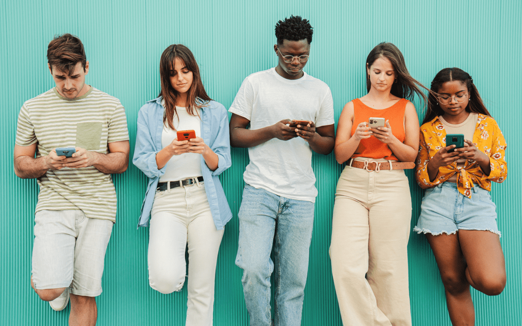Five young people of diverse backgrounds stand against a turquoise wall while looking at their mobile phones.