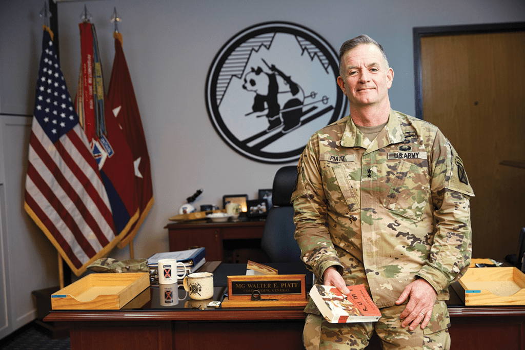 A military officer in camouflage uniform stands in an office with American and military flags in the background.