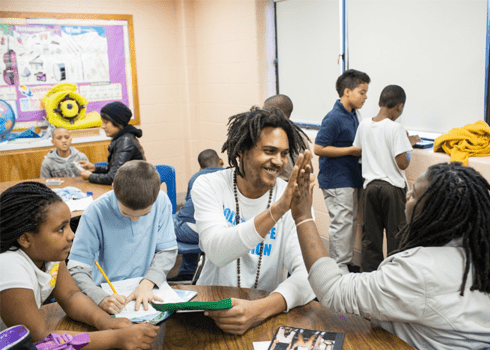 A teacher high-fives a student in a classroom, surrounded by other students engaged in various activities like writing and drawing. The room is lively with educational posters on the walls and school supplies on the desks.