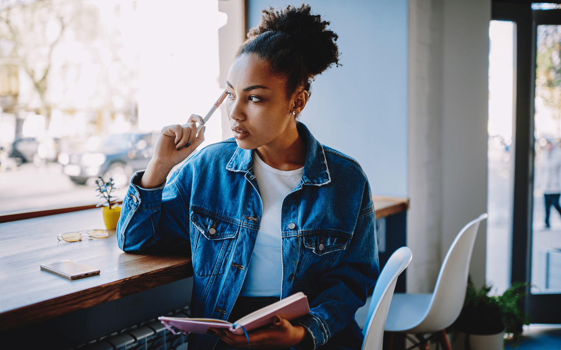 A black woman looks out of the window holding a book, with a pencil in her hand and has a thoughtful expression.